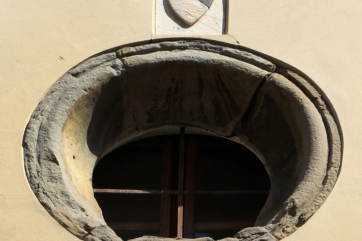 Et rundt stenstensvindue indsat i en lyspudset mur på Via Santa Monaca i Firenze, med et lille stenskjold over det